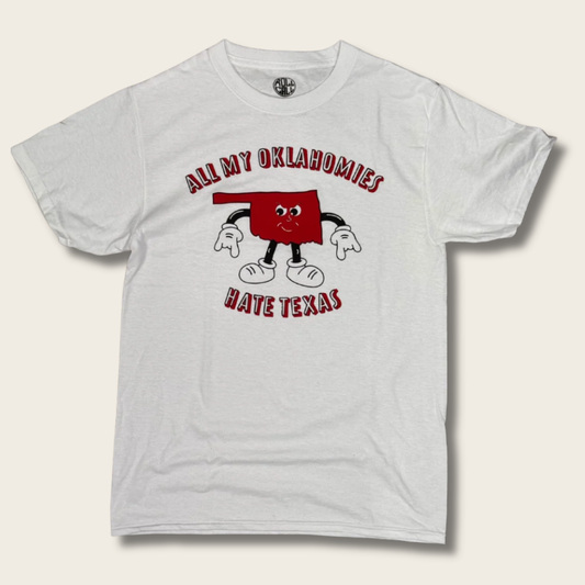 A white short-sleeve graphic t-shirt featuring a cartoon illustration of the state of Oklahoma with arms and legs, accompanied by the text 'ALL MY OKLAHOMIES HATE TEXAS' in red arched lettering.