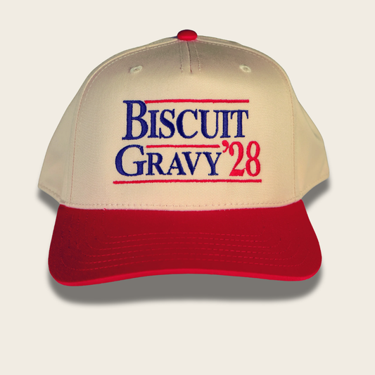 A baseball cap with a cream-colored crown and a red visor, featuring the embroidered text 'BISCUIT GRAVY '28' in blue and red on the front. The cap is shown against a light gray background with a soft drop shadow.