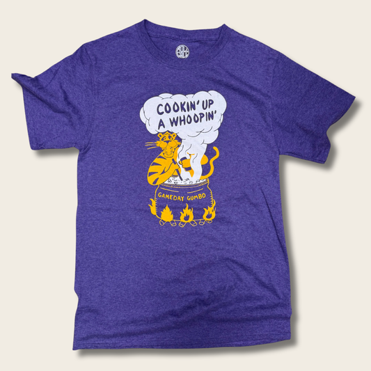 A heather purple short-sleeve t-shirt with a graphic of a yellow cat cooking in a pot and a speech bubble that reads 'COOKIN' UP A WHOOPIN''.