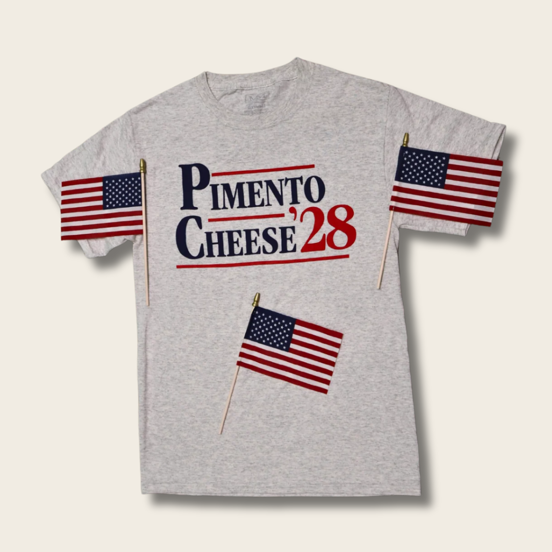 A light heather gray t-shirt featuring a 'PIMENTO CHEESE '28' graphic in navy and red on the chest, with American flags printed on both sleeves and a small American flag graphic below, shown against a light beige background.