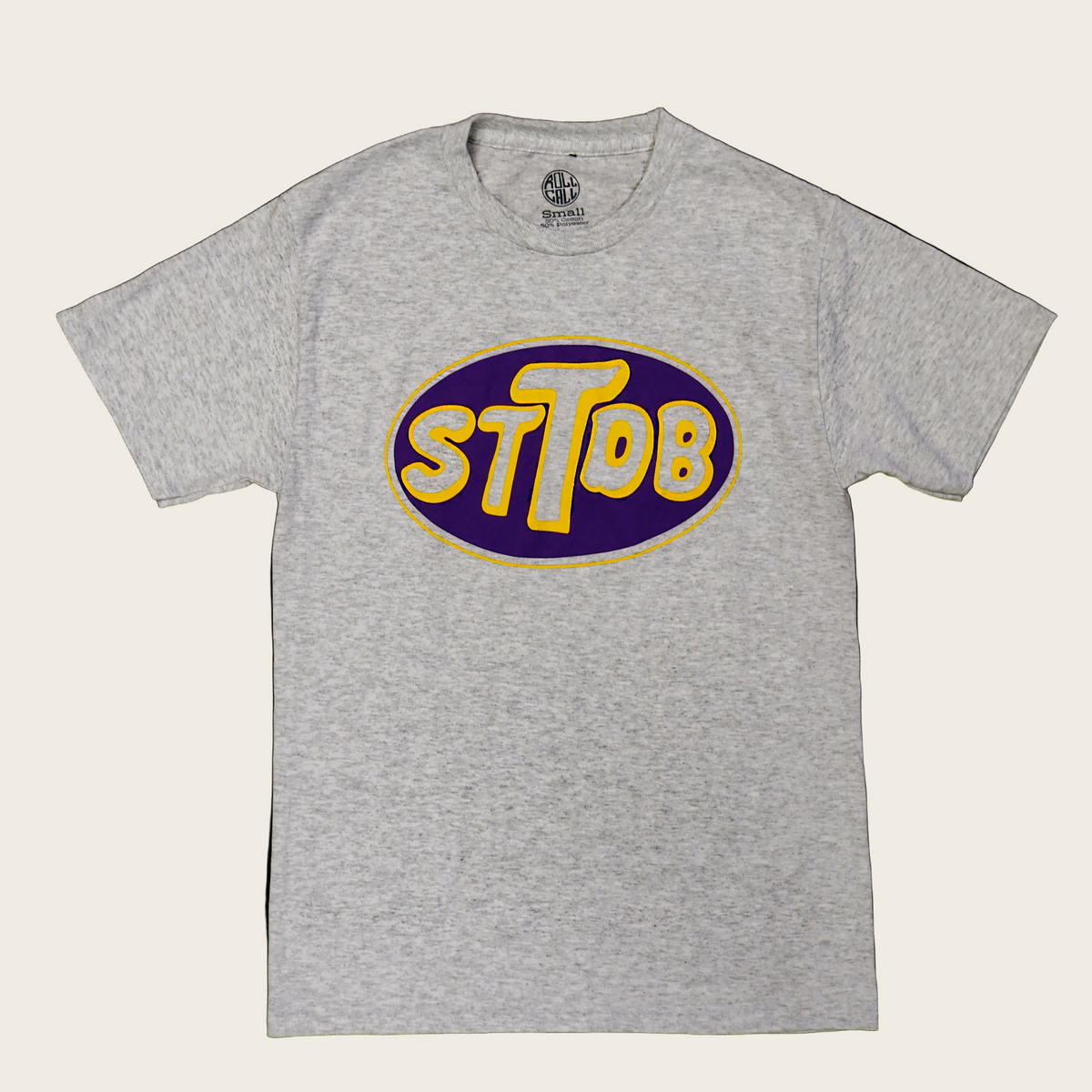 A heather gray short-sleeve t-shirt with a large oval graphic on the chest featuring the text 'STTDB' in yellow with a white outline on a dark purple background.