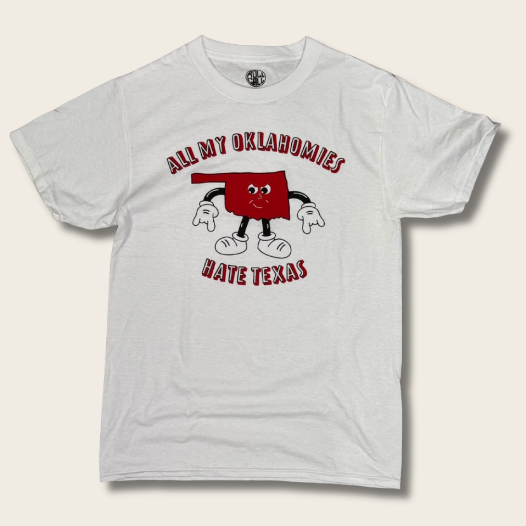 A white short-sleeve graphic t-shirt featuring a cartoon illustration of the state of Oklahoma with arms and legs, accompanied by the text 'ALL MY OKLAHOMIES HATE TEXAS' in red arched lettering.