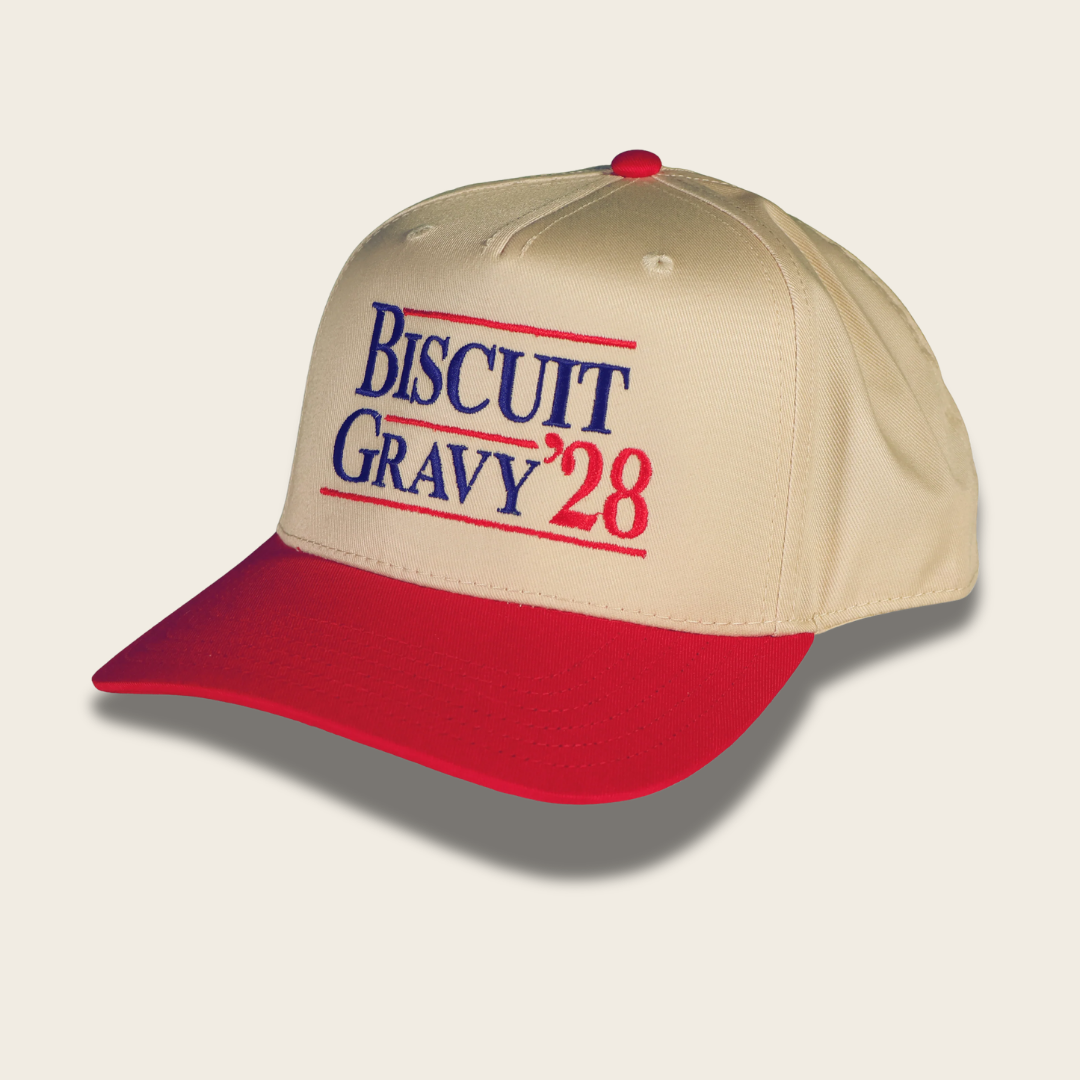 A beige baseball cap with a red curved brim, featuring embroidered text on the front that reads 'BISCUIT GRAVY '28' in blue and red lettering, shown on a light beige background.