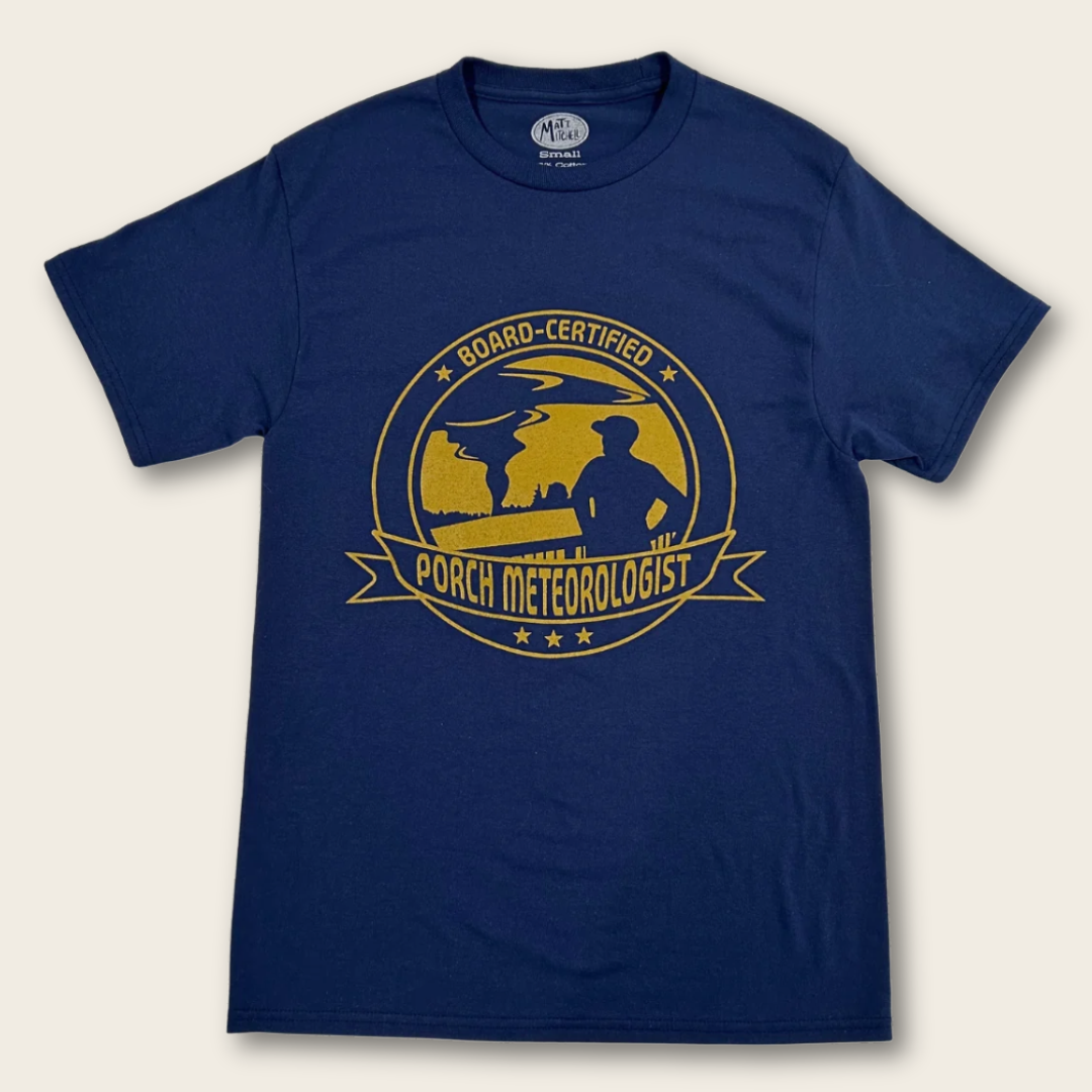 A navy blue short-sleeve t-shirt with a distressed yellow graphic on the chest that reads 'BOARD-CERTIFIED PORCH METEOROLOGIST' with a silhouette of a person on a porch watching a tornado in the distance.