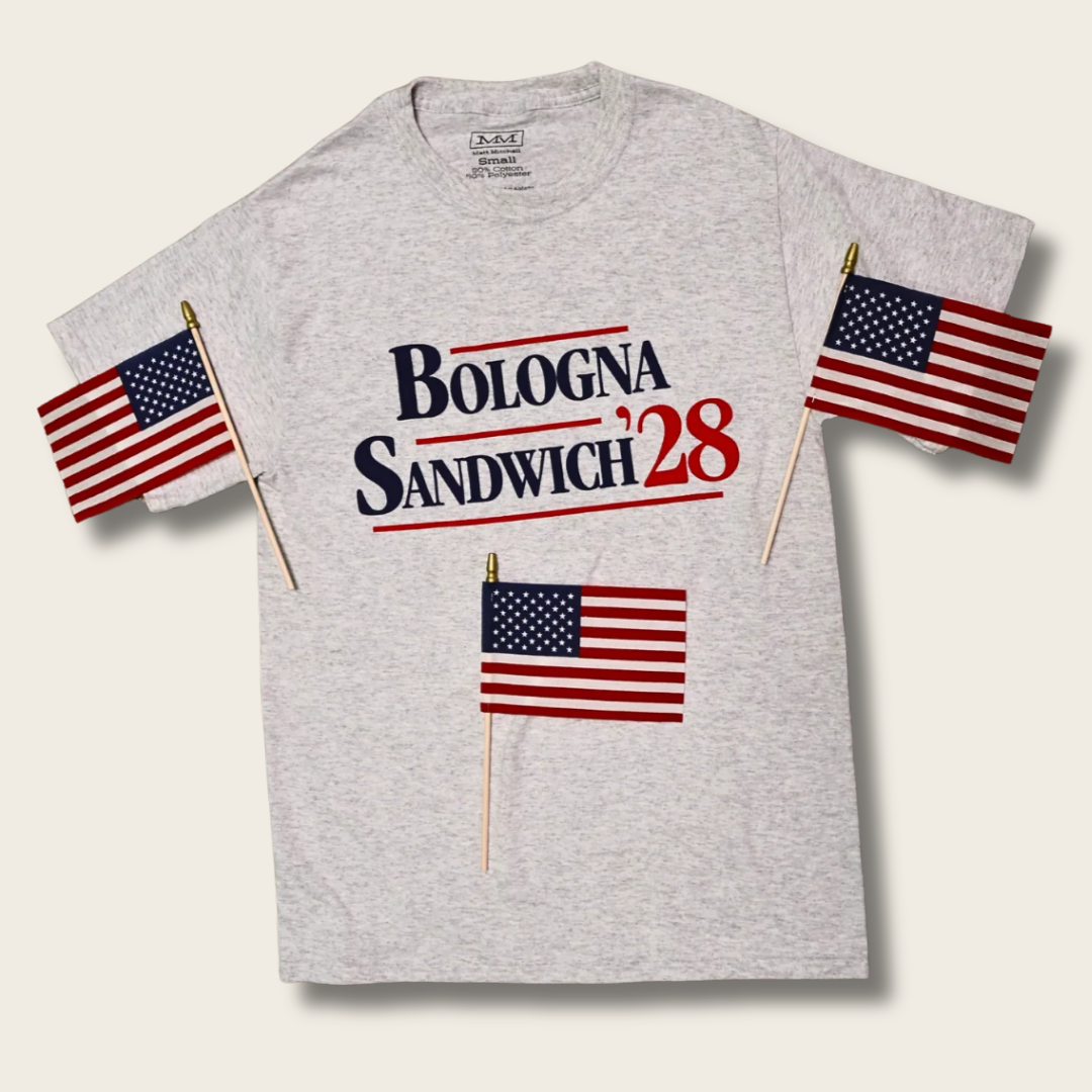 A light heather gray t-shirt featuring the text 'BOLOGNA SANDWICH '28' in navy and red lettering, styled with three miniature American flags on sticks, displayed against a light beige background.
