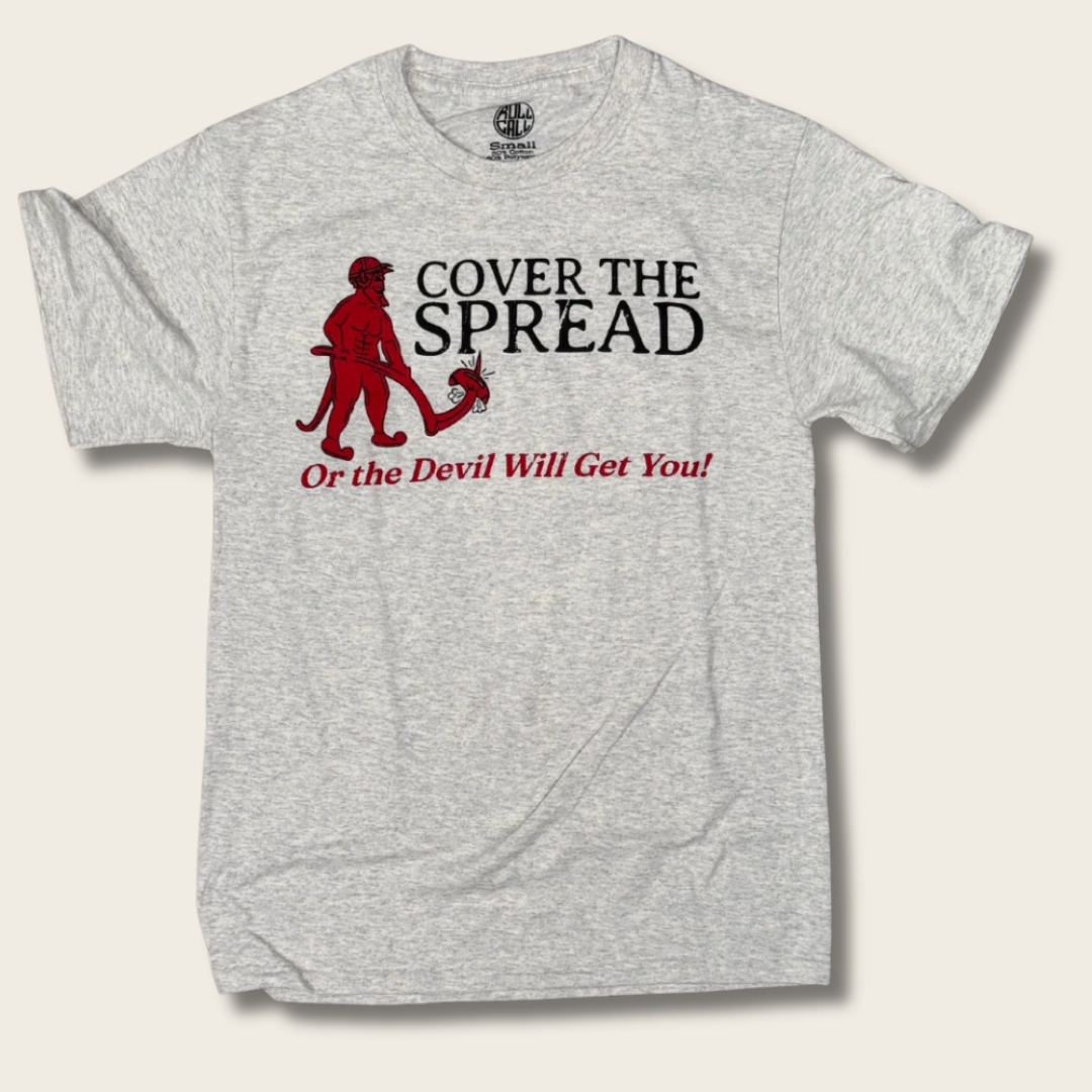 A light heather gray t-shirt with a graphic print featuring a red devil holding a book and the text 'COVER THE SPREAD Or the Devil Will Get You!' in black and red, displayed on a light beige background.