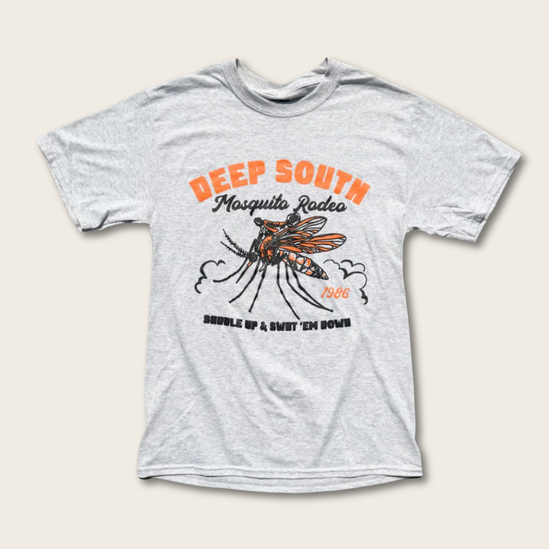 A gray shirt featuring a cowboy riding a large mosquito, with the text "DEEP SOUTH Mosquito Rodeo SADDLE UP & SWAT 'EM DOWN 1986" in orange and black text, all on a cream background.