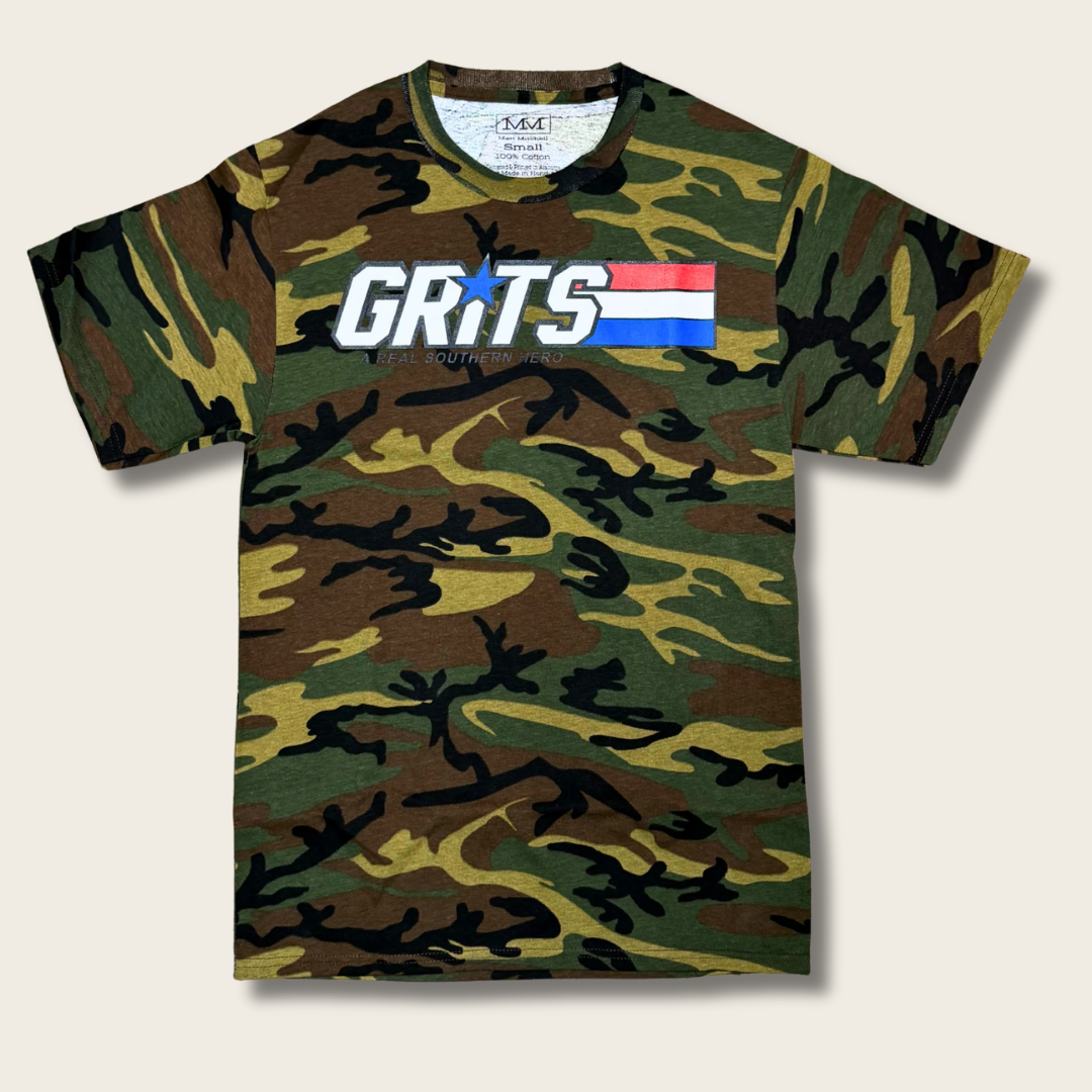 A short-sleeve crewneck t-shirt featuring a green, brown, and black camouflage pattern. The chest is printed with the word 'GRITS' in bold white letters, with a blue star replacing the 'I', and a small red, white, and blue flag graphic behind it.
