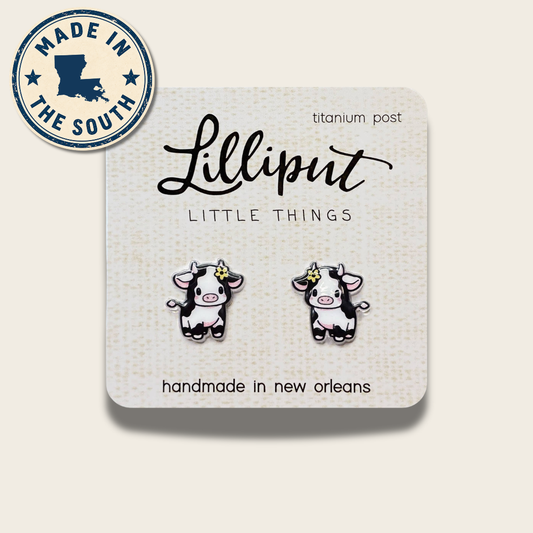 A pair of cartoon cow stud earrings on a white display card. The card reads 'Lilliput LITTLE THINGS' and 'handmade in new orleans,' with a circular 'MADE IN THE SOUTH' badge in the top left.