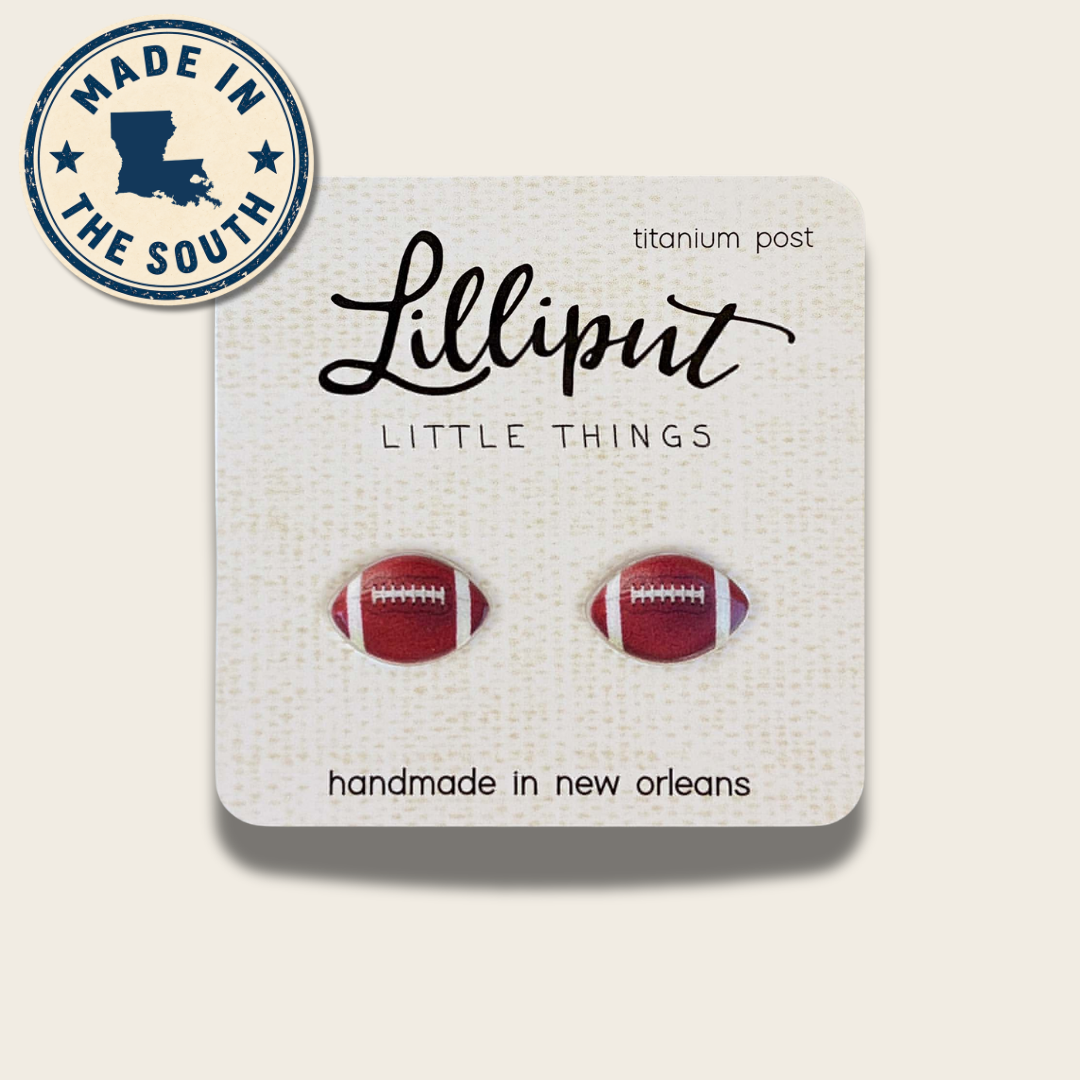 A pair of football-shaped stud earrings in red and white enamel, mounted on a white textured display card. The card features the brand name 'Lilliput' and the text 'LITTLE THINGS' and 'handmade in new orleans'.