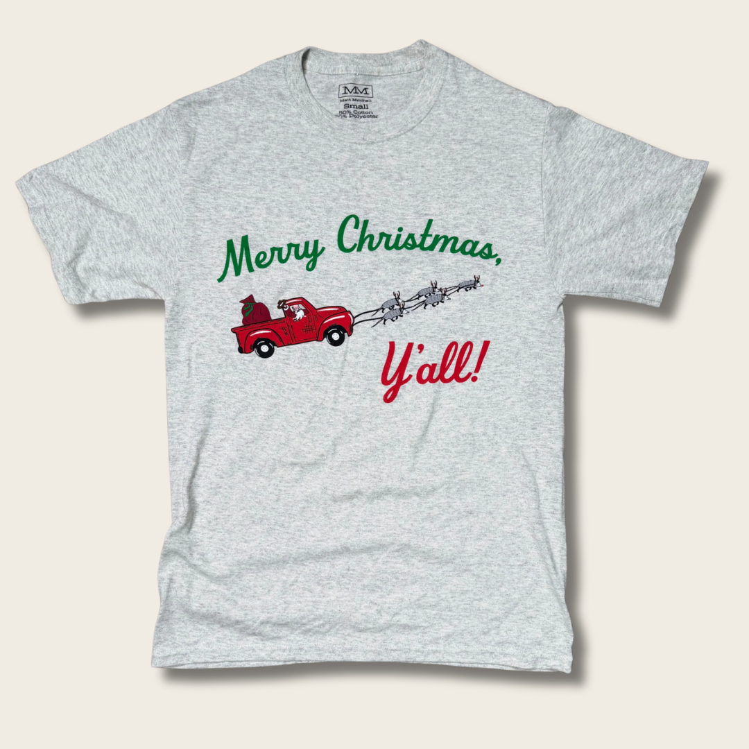 A light heather gray short-sleeve t-shirt with a Christmas graphic on the front. The graphic features a red pickup truck carrying a Christmas tree, with the text 'Merry Christmas, Y'all!' in green and red script.