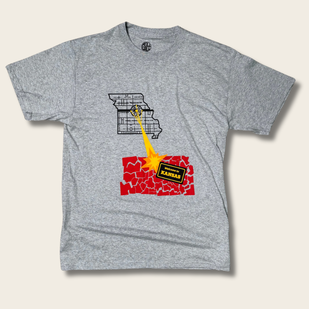 A heather gray short-sleeve t-shirt with a graphic print of Missouri and Kansas in a 'battle stance' facing each other, with a yellow explosion between them. The word 'KANSAS' is printed on a black banner in front of the red cracked Kansas graphic.