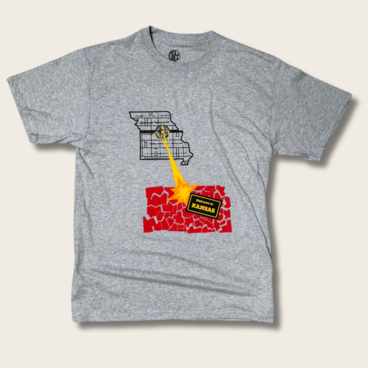 A heather gray short-sleeve t-shirt with a graphic print of Missouri and Kansas in a 'battle stance' facing each other, with a yellow explosion between them. The word 'KANSAS' is printed on a black banner in front of the red cracked Kansas graphic.