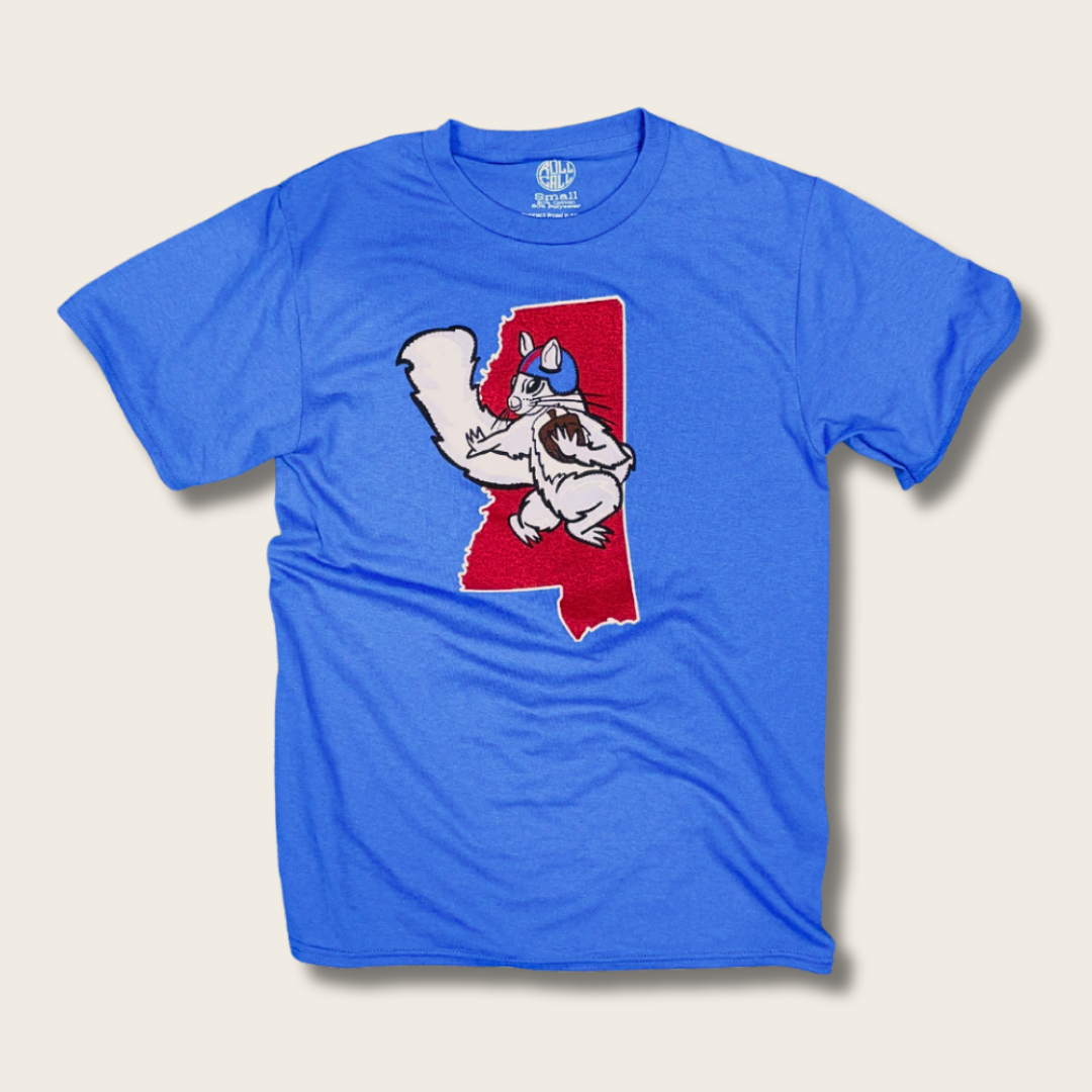 A blue short-sleeve crewneck t-shirt featuring a graphic of a white cartoon squirrel wearing a blue helmet, superimposed on a red map-shaped outline on the front. The shirt is displayed flat against a light beige background.