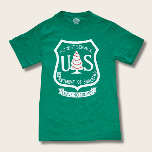A green short-sleeve crewneck t-shirt featuring a white graphic print of a Christmas tree cake, with the white text "FOREST SERVICE U.S. DEPARTMENT OF SNACKING" with bottom text in a white banner, "LEAVE NO CRUMBS" on a cream background.