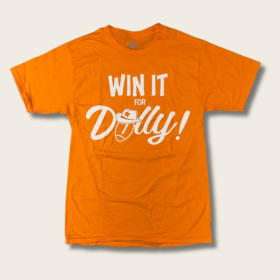 An orange short-sleeve t-shirt with a white graphic print reading 'WIN IT FOR Dolly!' with a football and cowboy hat illustration, displayed on a light beige background.